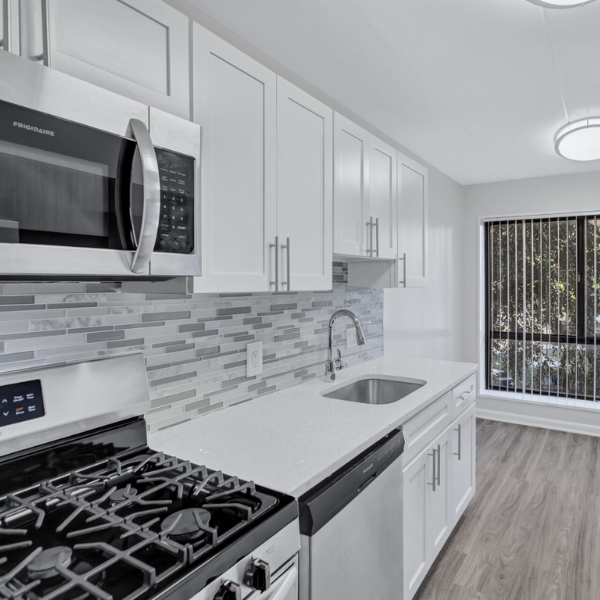 Kitchen Interior - CITYSIDE Huntington Metro A modern living room with a loft design features a metal staircase leading to an upper level. Two large windows with blinds allow natural light to fill the space. The room includes a gray couch, a coffee table with a glass fireplace, and a floor lamp.